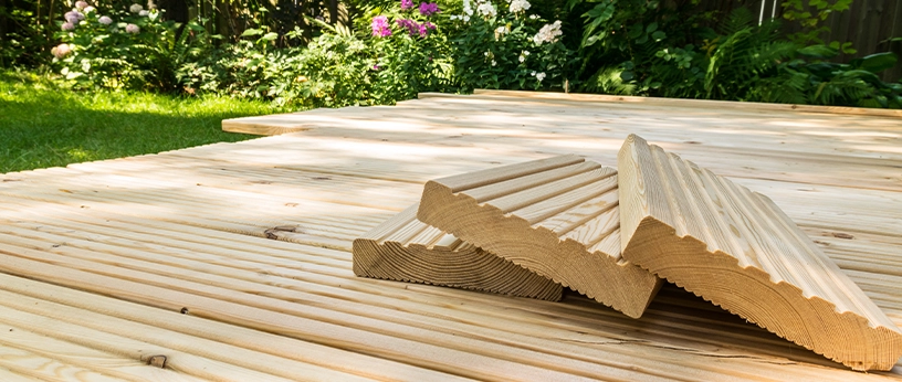 Terrassenbau mit hellen Lärchenholzdielen, gestapelte Riffelbretter im Vordergrund, umgeben von grünem Garten.