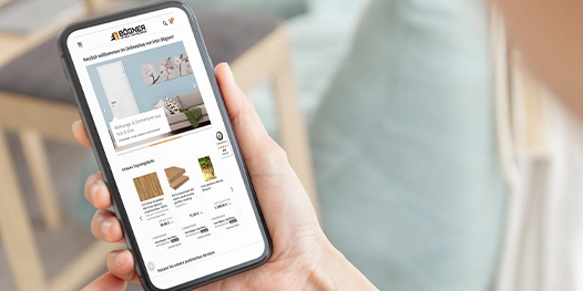 Hand hält Smartphone mit geöffneter Website, die Produkte aus dem Bereich Wohnen und Einrichten zeigt, darunter Möbel und Wohnaccessoires.