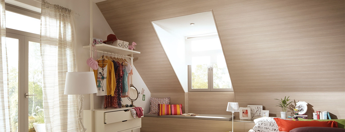 Helles Dachzimmer mit Holzverkleidung, Fensterfront, offener Kleiderstange und gemütlicher Sitzecke mit Kissen.