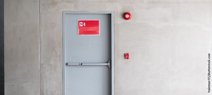 Graue Brandschutztür mit Fluchtwegsymbol und roter Alarmglocke.