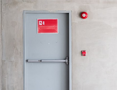 Graue Brandschutztür mit Fluchtwegsymbol und roter Alarmglocke.