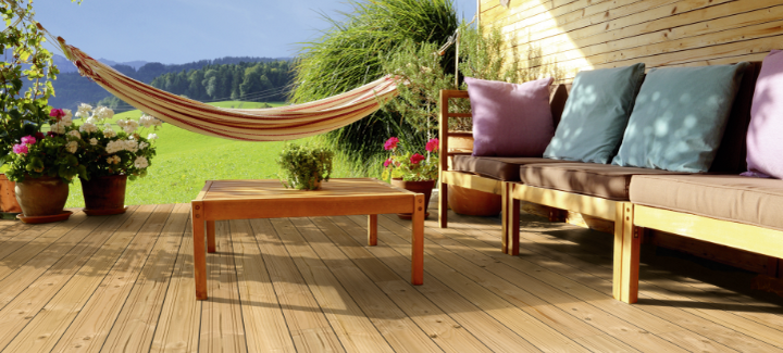 Gemütliche Holzterrasse mit Douglasien-Dielen, Lounge-Sofa mit Kissen, Holztisch, Hängematte und blühenden Pflanzen vor idyllischer Landschaft.