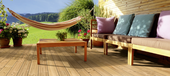 Gemütliche Holzterrasse mit Douglasien-Dielen, Lounge-Sofa mit Kissen, Holztisch, Hängematte und blühenden Pflanzen vor idyllischer Landschaft.